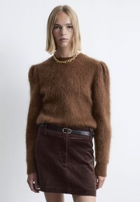 Brauner, flauschiger Pullover mit Rundhalsausschnitt und Puffärmeln, kombiniert mit einem braunen Cordrock mit einem taillierten Gürtel. Goldkettchen-Halskette.