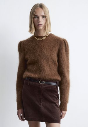 Brauner, flauschiger Pullover mit Rundhalsausschnitt und Puffärmeln, kombiniert mit einem braunen Cordrock mit einem taillierten Gürtel. Goldkettchen-Halskette.