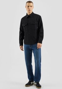 Czarna koszula zapinana na guziki z dwoma kieszeniami na piersi, zestawiona z niebieskimi dżinsami i czarnymi trampkami. Gładka tkanina o strukturalnym kroju.