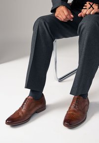 Chaussures habillées en cuir marron dotées d'un motif brogue avec des perforations, accompagnées d'un pantalon gris foncé à motif et de chaussettes bleu marine.