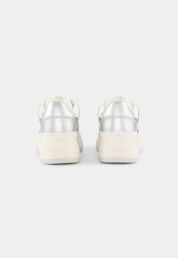 Białe sneakersy z srebrnymi akcentami, charakteryzujące się gładką skórzaną powierzchnią, grubymi białymi podeszwami i brandowanym tyłem. Minimalistyczny design ogólnie.