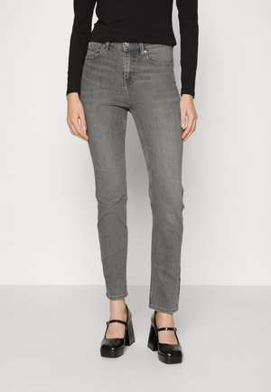 Vrouw draagt grijze skinny jeans, zwarte top met lange mouwen en zwarte Mary Jane schoenen met hoge hakken, staand tegen een witte achtergrond.