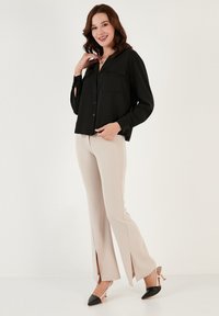 Camicia nera con bottoni e due taschini sul petto, abbinata a pantaloni beige con orlo spaccato e tacchi neri e beige. Tessuto liscio e leggero.