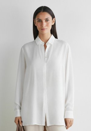 Junge Frau mit glattem, dunklem Haar, die ein weißes, langärmliges Hemd mit Knöpfen trägt und einen braunen Gegenstand vor einem schlichten Hintergrund hält.