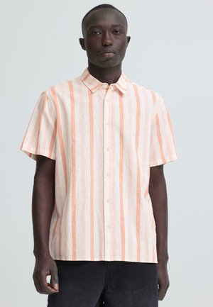 Mężczyzna stoi przodem, ubrany w koszulę na guziki z krótkim rękawem w pionowe różowo-białe paski oraz czarne spodnie.