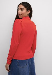 Pullover rosso con colletto alto, tessuto testurizzato e dettaglio arricciato sulle spalle, abbinato a una gonna di jeans blu.