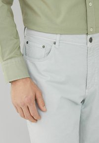 Hellgrünes Hemd mit langen Ärmeln, kombiniert mit hellblauen Hosen, die eine Fronttasche und metallische Akzente aufweisen.