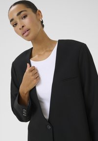 Čierny blazer s jedinečným otvoreným dizajnom, s jedným gombíkom na zapínanie a jemným detailom vrecka. Modelka má pod ním biely top.