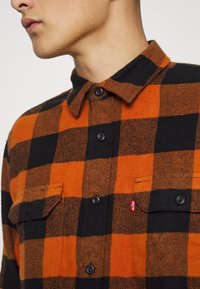 Chemise en flanelle à carreaux noirs et orange avec un design à boutons, deux poches avant et le logo Levi's sur la couture de la poche gauche.