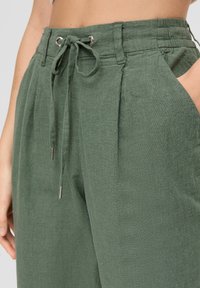 Grüne Leinenhose mit einem Kordelzugbund, seitlichen Taschen und Falten. Glatte Textur und metallische Hardware-Akzente am Kordelzug.
