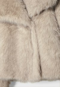 Manteau en fausse fourrure beige clair, doux, à texture dense et duveteuse, montrant le col et les panneaux avant en gros plan.