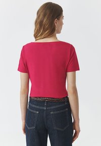 Top court à manches courtes en tissu rose vif avec un décolleté carré, associé à un jean bleu foncé taille haute et une ceinture tressée marron.