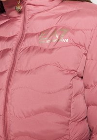 Růžová prošívaná péřová bunda se zapínáním na zip a zlatými logy. Měkká, lehká látka s jemným leskem.