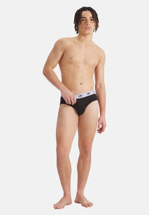 Jonge man met sproeten en dreadlocks, staand op blote voeten, dragend een zwarte slip met een witte tailleband met een zwart trefoil-logo.