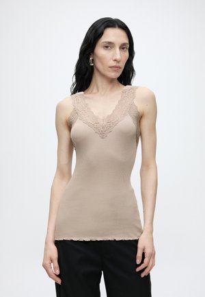 Mujer con cabello largo y negro que lleva una camiseta sin mangas beige con encaje y pantalones negros, de pie frente a un fondo claro y liso.