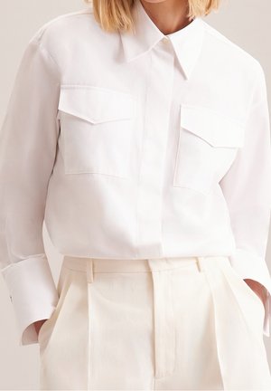 Mujer con camisa blanca de manga larga abotonada con bolsillos en el pecho con solapa, metida en pantalones plisados crema de tiro alto.