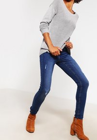Femme portant un pull en maille gris, un jean skinny bleu foncé légèrement usé et des bottines en suède marron, posant jambes écartées.
