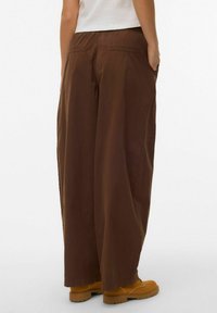 Bruine broek met wijde pijpen, een elastische tailleband, zijzakken en een gladde textuur. Gecombineerd met geel schoeisel, gezien van achteren.