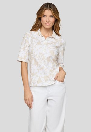 Woman with light brown hair wearing a beige floral short-sleeve polo shirt and white pants standing with one hand in her pocket.