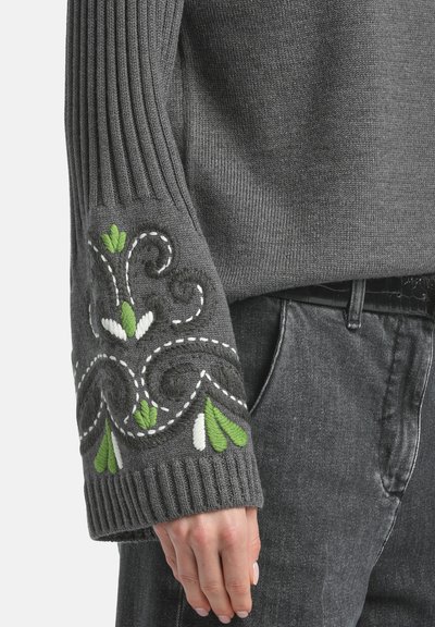 Grauer Strickpullover mit gerippten Bündchen und grüner sowie weißer Blumenstickerei, in lockerer Passform. Kombiniert mit schwarzen Jeans.