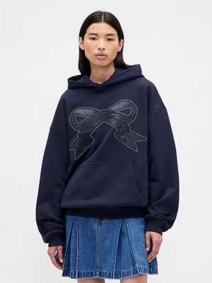 Sudadera azul-marinho com um grande laço em faux leather preto, realçado por tachas brancas. Apresenta um bolso canguru e um ajuste descontraído, combinado com uma saia de ganga.