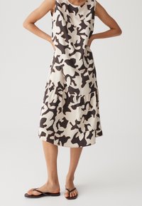Femme portant une robe mi-longue sans manches à motif abstrait noir et blanc avec un détail volanté et des sandales tongs noires.