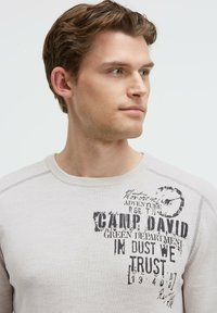 Hellgrauer Baumwollpullover mit Rundhalsausschnitt, mit schwarzen Druckgrafiken. Der Text beinhaltet "CAMP DAVID" und "IN DUST WE TRUST".