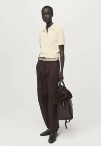 Donna che indossa un top a maglia corto color crema, pantaloni marrone scuro, stivali neri, mentre tiene una grande borsa marrone con dettagli in pelliccia.