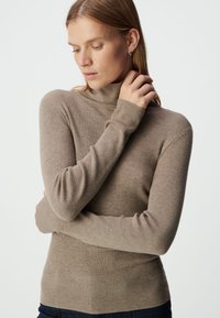 Beige geribbelde coltrui met lange mouwen, met een getailleerd silhouet en een zachte, gestructureerde stof. Model houdt haar arm voorzichtig vast.