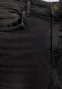 Nærbillede af en frontlomme på sorte denim jeans, bælteslynge og metalknap med indgraveret mærkenavn.