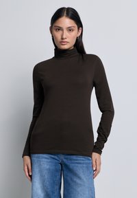 Jonge vrouw met lang, donker haar, gekleed in een zwarte coltrui en blauwe spijkerbroek, tegen een effen lichte achtergrond.