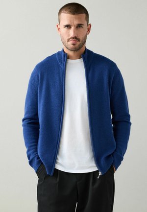 Blauer Reißverschluss-Cardigan aus weichem Material, mit hohem Kragen, gerippten Bündchen und Seitentaschen, über einem weißen Hemd getragen.