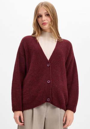 Giovane donna che indossa un cardigan di maglia bordeaux con bottoni sopra una maglietta beige a coste con collo alto e pantaloni grigio chiaro su uno sfondo semplice.