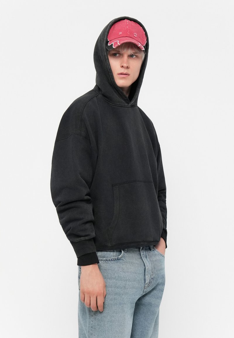 Felpa nera con tasca a marsupio, tessuto testurizzato e polsini a coste; abbinata a un cappellino rosa con dettagli di cucitura.