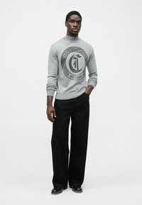 Sweatshirt gri cu guler rotund și logo grafic negru cu model de piele de șarpe, asociat cu pantaloni negri cu picioare largi și încălțăminte.