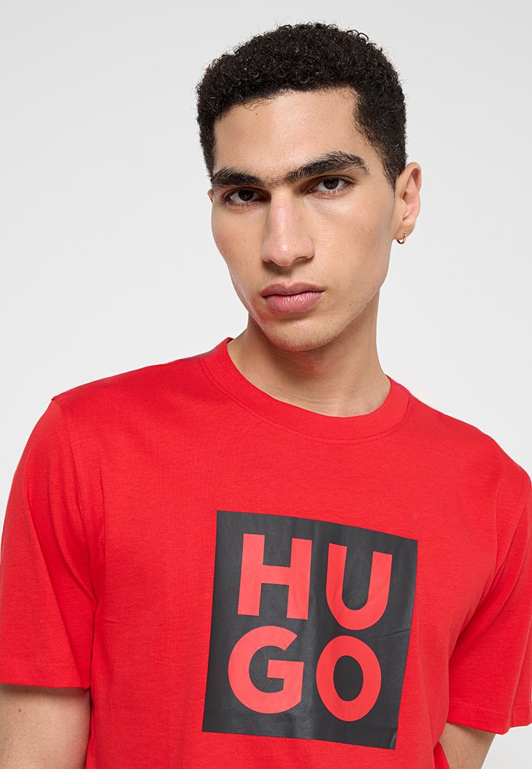 Camiseta de algodón roja con un gran gráfico cuadrado negro que lleva la palabra "HUGO" en tipografía negrita roja y negra. Con mangas cortas y cuello redondo.