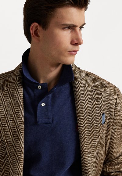 Brun herringbone-blazer over en marineblå polo med to knapper, som har en sydd logo og en blå lomme detalj.