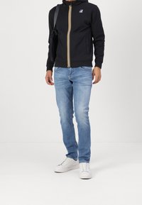 Uomo che indossa jeans slim blu chiaro, sneakers bianche e una felpa nera con zip, dotata di zip a righe e logo sul petto, mentre porta una borsa nera.