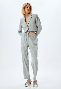 Gekürzter grauer Blazer mit einem einzelnen Knopf und Fronttasche, kombiniert mit passenden Stoffhosen mit elastischem Bund und metallischen spitzen Absätzen.