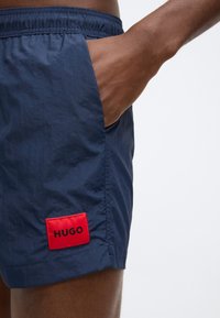 Námořnicky modré plavkové šortky z lehké tkaniny mají pružný pas, boční kapsy a červenou nášivku s logem "HUGO" na lemu.