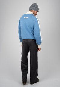 Homme portant un pull bleu à rayures blanches, un pantalon foncé, un bonnet gris en tricot et des chaussures noires, debout et regardant vers le bas sur un fond gris.