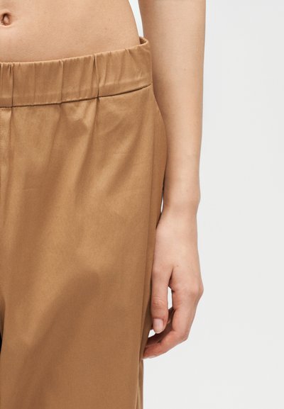 Κοντινό πλάνο της μεσαίας περιοχής ενός ατόμου που δείχνει μπεζ ελαστικό παντελόνι μέσης και ένα χαλαρό δεξί χέρι δίπλα στο ισχίο.