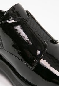 Chaussure en cuir verni noir avec une large bande élastique sur le dessus, une semelle mate contrastante et un design à bout arrondi. Finition lisse et brillante.