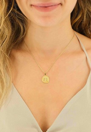 Collier pendentif en or avec un charme circulaire texturé orné d'un motif, suspendu à une fine chaîne en or, porté sur un haut de couleur claire.