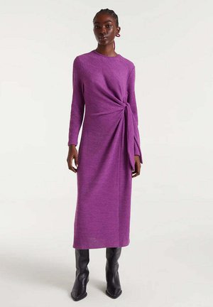 Mujer vestida con un vestido midi de manga larga morado con detalle de nudo lateral, combinado con botas negras hasta la rodilla sobre un fondo claro.