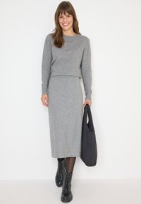 Femme portant un pull gris à manches longues et une jupe assortie, marchant avec un sac fourre-tout noir, chaussée de bottes noires et souriant.