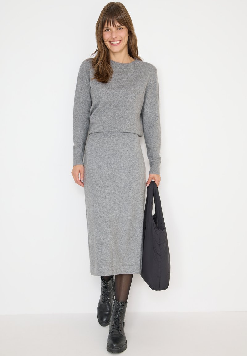 Femme portant un pull gris à manches longues et une jupe assortie, marchant avec un sac fourre-tout noir, chaussée de bottes noires et souriant.