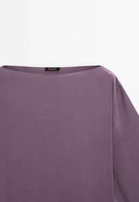 Haut ample à manches courtes en tissu violet clair. Présente un large col et une texture douce et lisse. Étiquette de la marque visible.