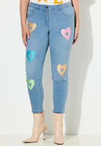 Vaqueros pitillo azul claro con aplicaciones de corazones de lentejuelas en colores dorado, rosa, verde y teal. Diseñados con una cintura de talle medio.