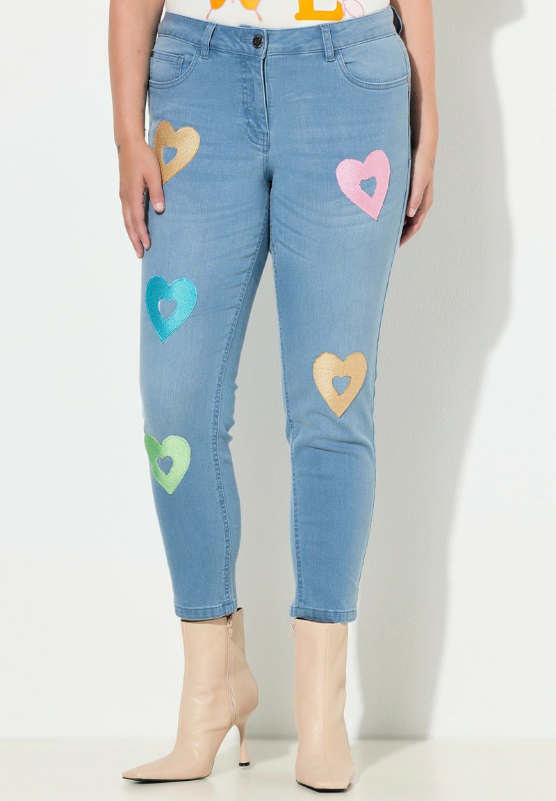 Vaqueros pitillo azul claro con aplicaciones de corazones de lentejuelas en colores dorado, rosa, verde y teal. Diseñados con una cintura de talle medio.